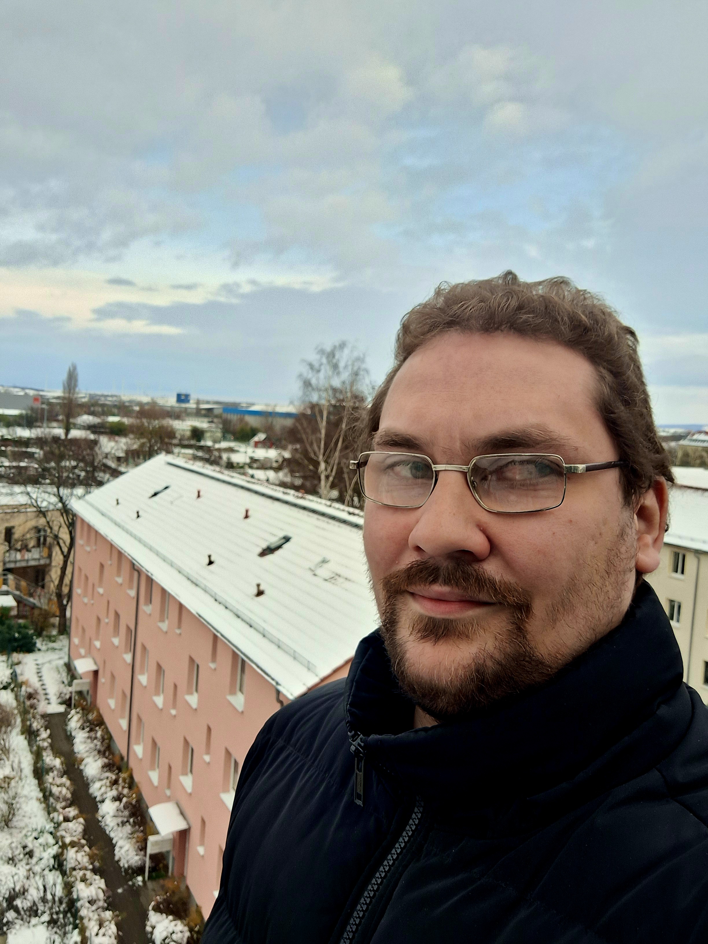 Ich stehe auf einer Terasse wärend ich leicht lächel. Hintermir ist die verschneite Johannesvorstadt in Erfurt zu sehen.